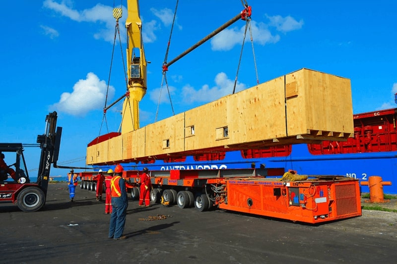 6 men in hardhats, a large crane & forklift unloading & loading a 50-foot wooden crate at seaport